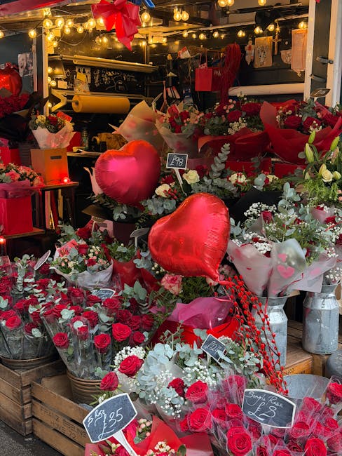 A floral display featuring an arrangement of fresh red roses, white filler flowers, and greenery, wrapped in pink and white paper with heart-shaped balloons integrated into the bouquet. The display is