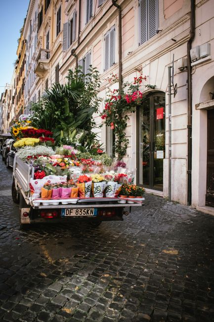 A delivery truck parked on a cobblestone street in front of a cream-colored building with tall, shuttered windows and an arched doorway. The truck bed is filled with an assortment of fresh floral arra