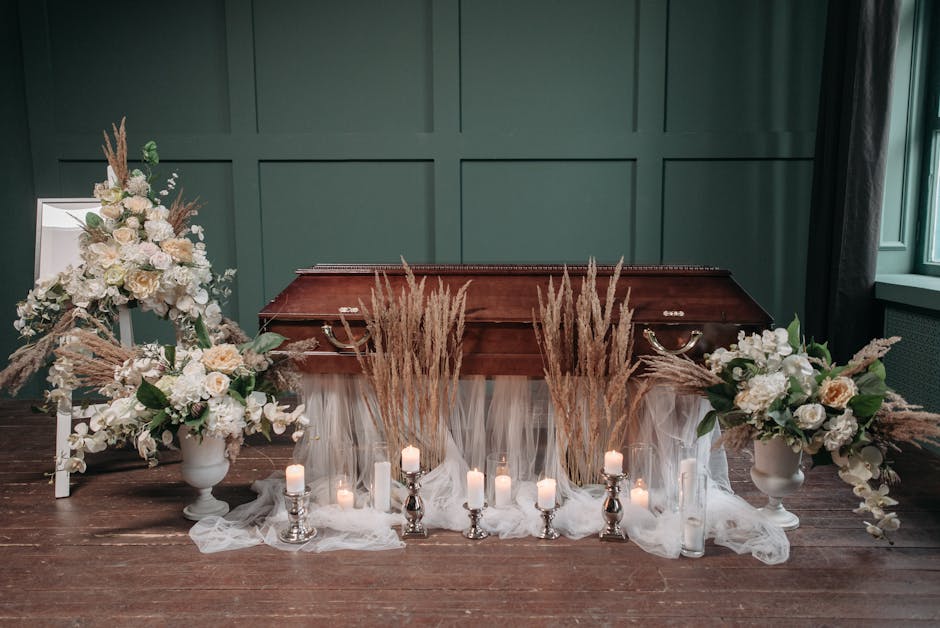 A funeral floral arrangement featuring two large white vases filled with ivory roses, white hydrangeas, and soft green foliage, complemented by cascading white and blush flowers. Tall, dried beige gra