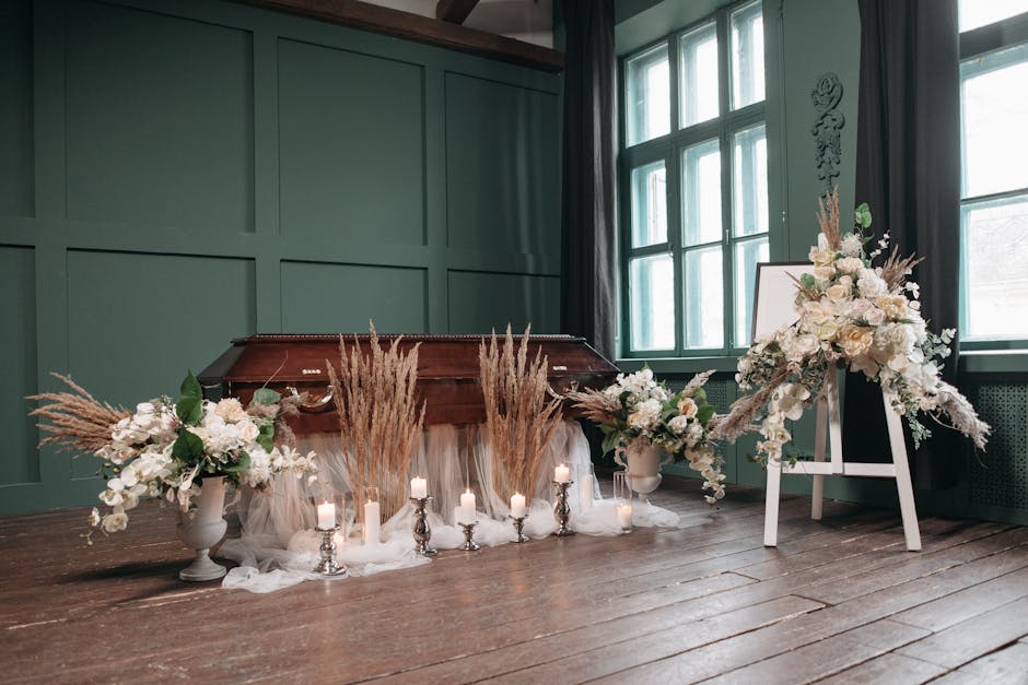 An elegant floral arrangement consisting of white roses, orchids, and soft pastel flowers in a white ceramic vase, displayed on a wooden floor near a window. The bouquet features lush greenery and is 