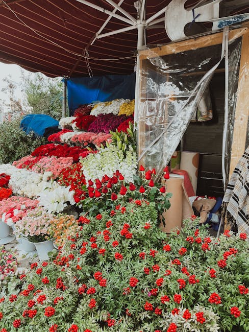 A young woman with long dark hair, wearing a white t-shirt and a black apron, is inside a florist shop, holding a glass vase filled with yellow tulips and some light pink and white flowers. The floral