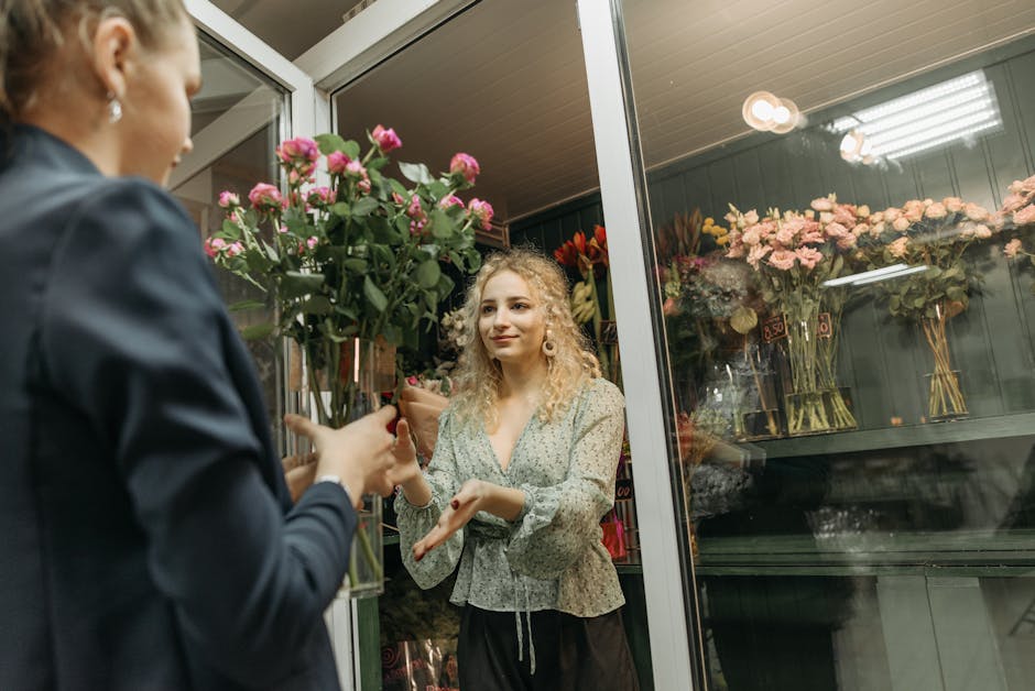 A young woman with long dark hair, wearing a white sleeveless top and beige trousers, is smiling and waving inside a florist shop filled with a variety of fresh flowers. She stands behind a counter th