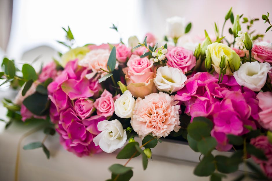 A young woman with light brown hair tied back, wearing a light grey apron over a blue shirt, is holding a fresh floral bouquet wrapped in pink and purple tissue paper. The bouquet includes various flo