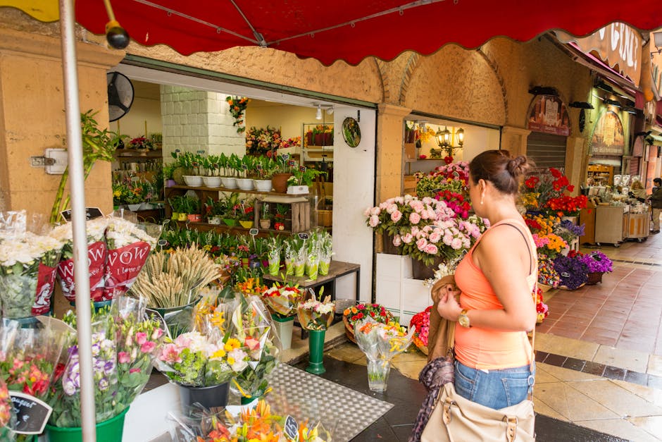 A woman stands in front of a florist shop with open white doors, browsing a colorful display of fresh flowers arranged in bouquets and potted plants. She holds a small bouquet in her hands and is dres