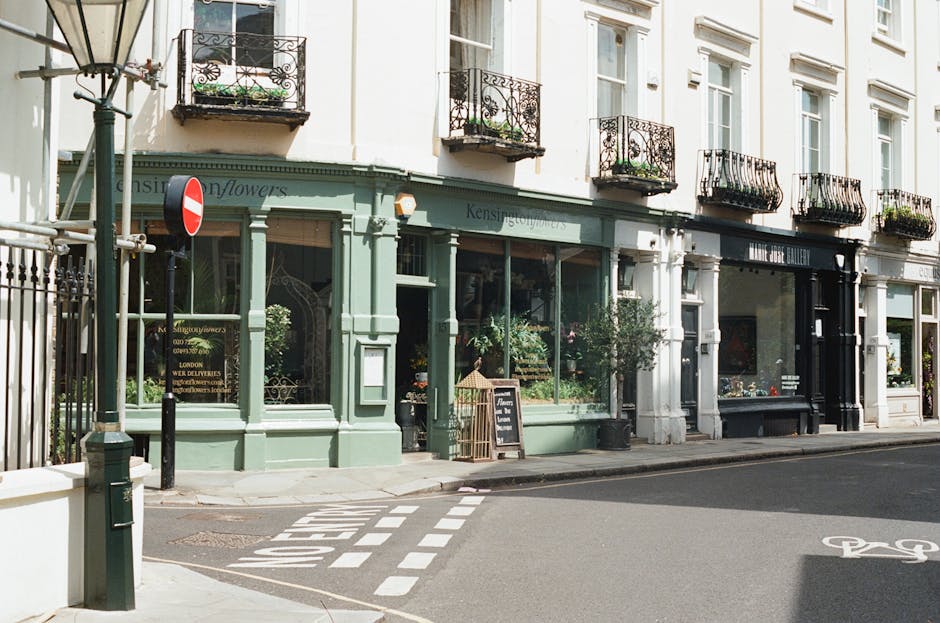 A floral boutique situated on the ground floor of a corner building with classical white window frames on a brick facade, featuring a large glass display window and a black door. Outside, a decorative