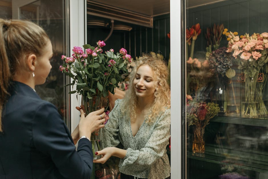 A florist from floristmaidavale.co.uk is delivering a bouquet of fresh pink roses with green foliage, wrapped in clear cellophane, to a smiling woman with curly blonde hair wearing a patterned blouse.