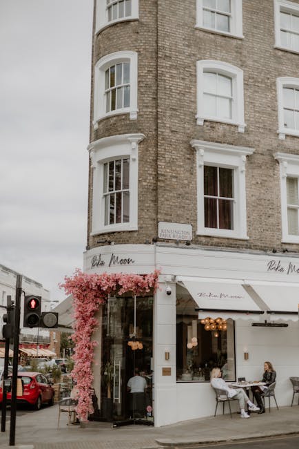 The storefront of a flower shop painted in a soft, pastel green shade with large display windows showcasing fresh floral arrangements and greenery inside. The shop is situated along a quiet street wit