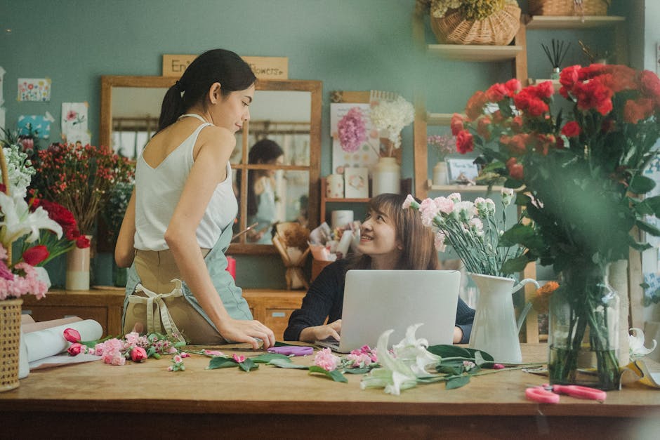 A young female florist with dark hair tied back, wearing a white sleeveless top and beige apron, standing in a flower shop surrounded by various fresh flowers, including pink carnations, white lilies,