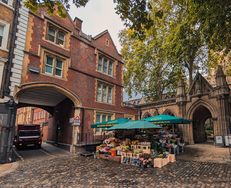 A flower stall situated on a cobblestone street near an arched stone gateway and a red brick building with multiple windows. The stall features a green umbrella with the website holbornflorist.co.uk, 