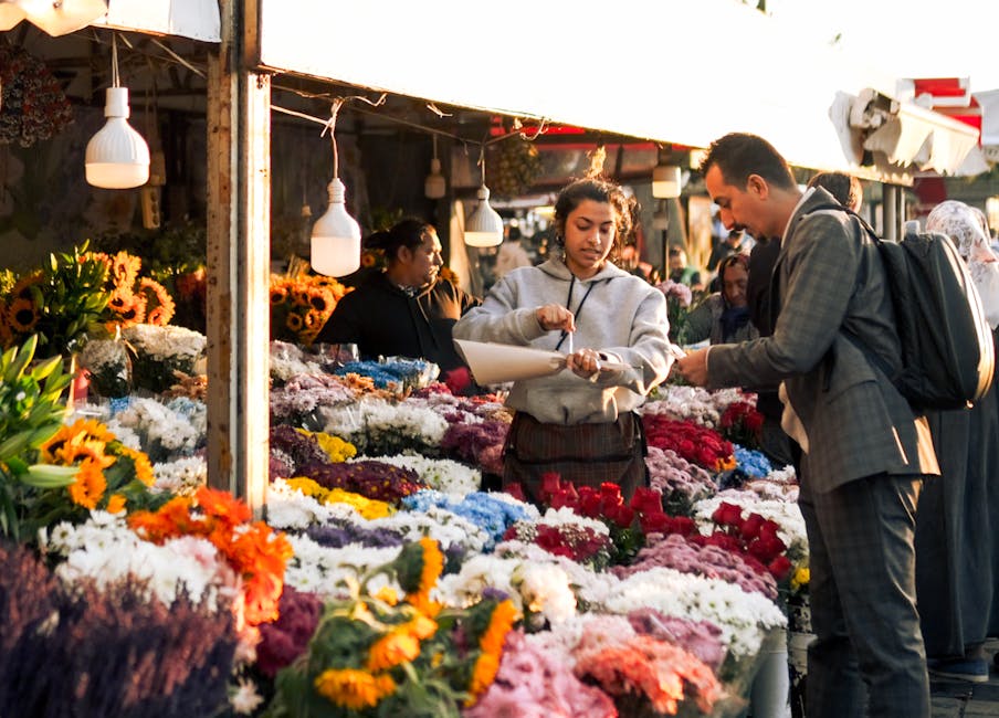 A vibrant display of floral arrangements and bouquets featuring red roses, white flowers, and greenery, displayed in metal buckets and wooden crates at a flower stall. The setup includes shiny red hea