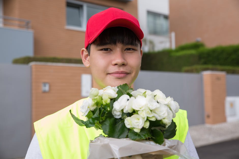 A young male delivery person wearing a red cap and a high-visibility yellow vest is holding a bouquet of white roses wrapped in white paper, with lush green leaves visible, in front of modern resident