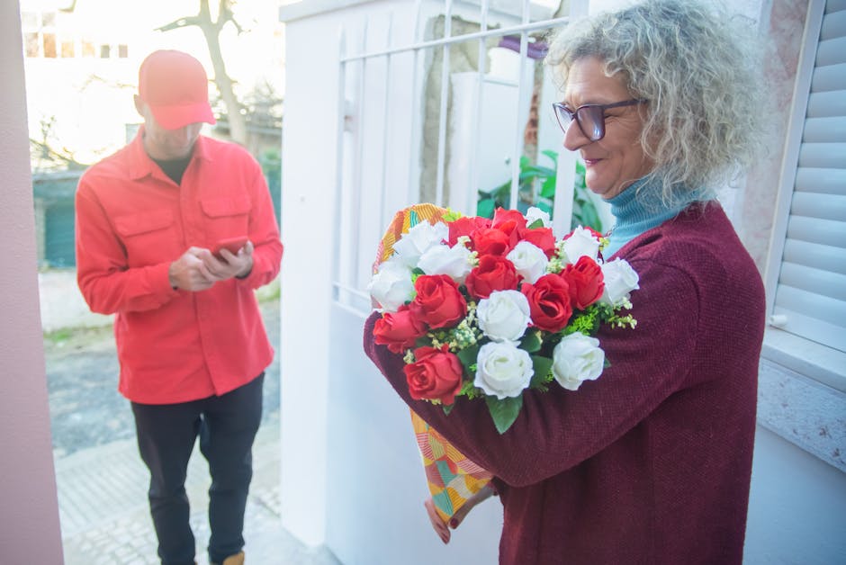A mature woman with curly gray hair and glasses is holding a vibrant bouquet of fresh red and white roses wrapped in colorful paper, standing near a white window frame and smiling gently. The bouquet 