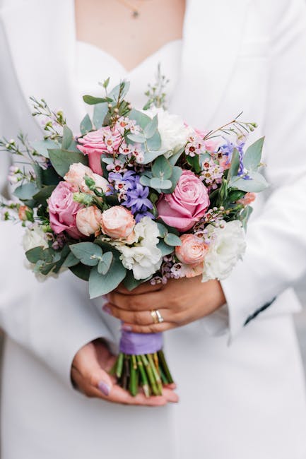 A person dressed in a white blazer holds a bouquet of fresh flowers, featuring soft pink roses, light purple lilacs, white lisianthus, and sprigs of eucalyptus with round, gray-green leaves. The bouqu