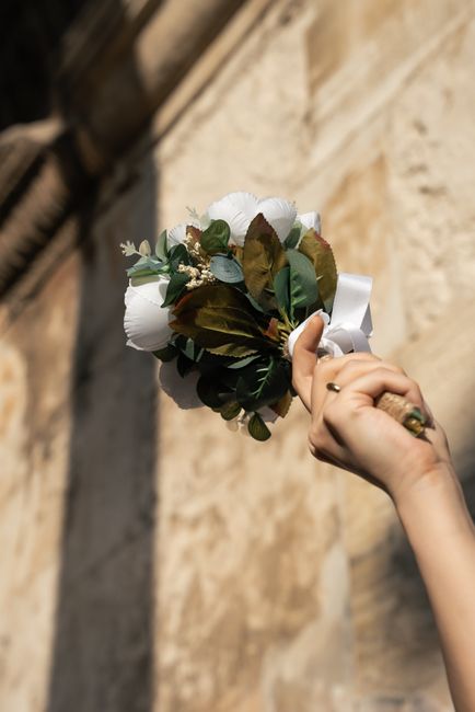 A close-up of a person's hand holding a small, elegant bouquet of fresh white roses and trailing greenery, accented with hints of brown and green leaves, tied together with a white satin ribbon. The p