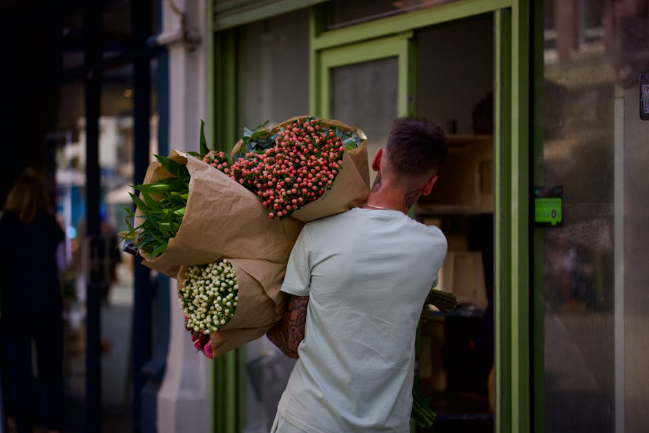 The image shows the exterior of a charming flower shop with a light green façade and large glass windows displaying floral arrangements inside. The storefront features a small outdoor chalkboard sign