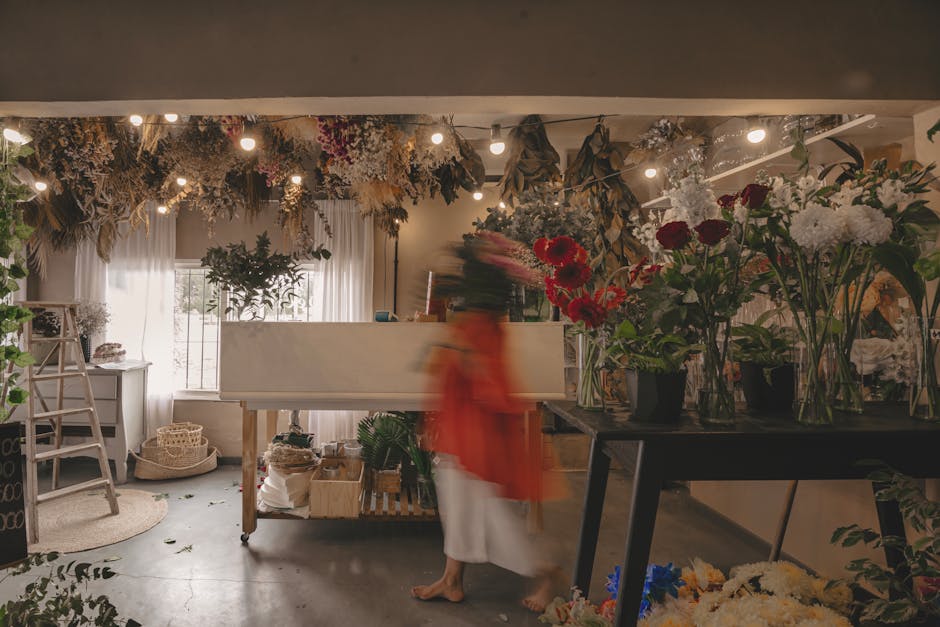 Inside a floral shop with natural daylight filtering through white curtains, displaying an arrangement of fresh flowers on a black table, including deep red roses, white and pink hydrangeas, and sprig